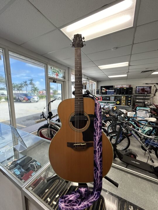 Takamine GN20-NS Nex acoustic guitar with natural finish displayed at Dixie Pawn, featuring a purple lightning-patterned strap.