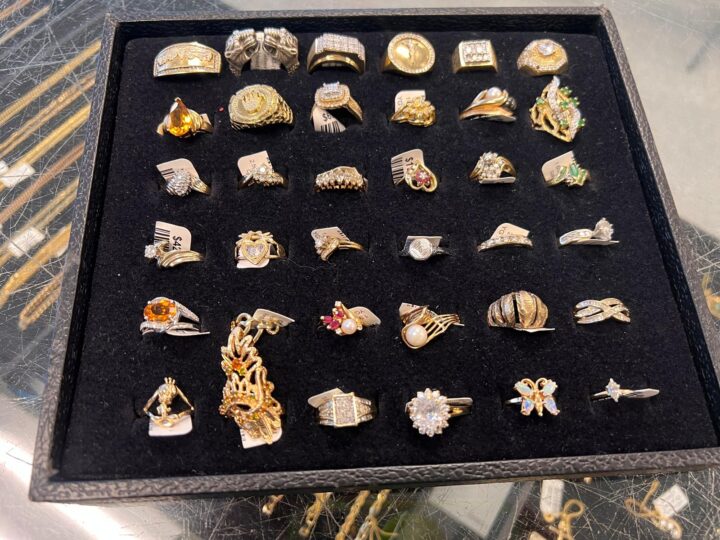 A display case featuring an assortment of gold and gemstone rings in various designs and styles, including intricate patterns, vibrant gemstones, and elegant settings, available for sale at Dixie Pawn in Hollywood, Florida.