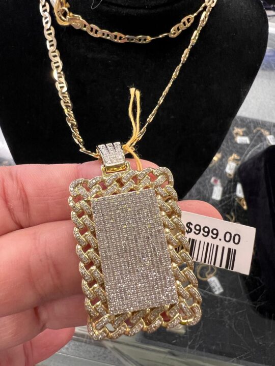 A close-up of a gold pendant densely set with diamonds, framed by a gold chain, priced at 9, held in hand at Dixie Pawn in Hollywood, FL.