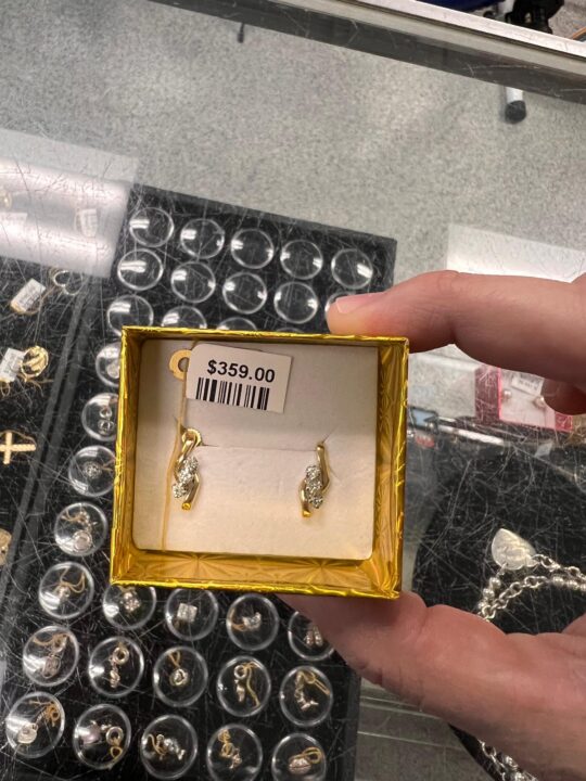 A person holding a gold box with a pair of gold earrings featuring diamond accents, priced at 9.00, set against a background of various jewelry items at Dixie Pawn in Hollywood, FL.