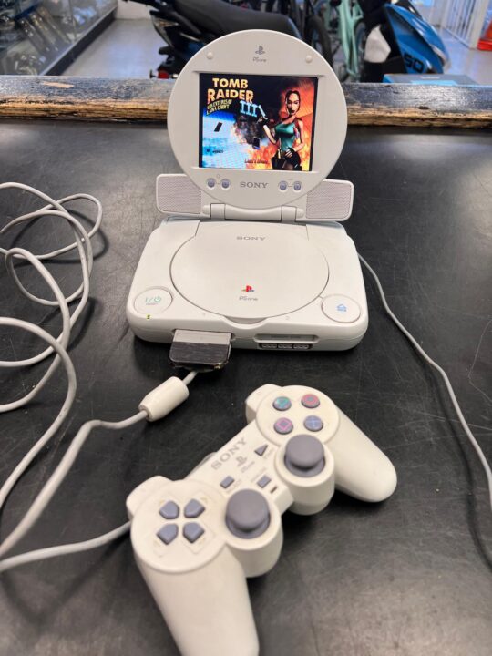 A Sony PSone with an attached screen displaying the game Tomb Raider III, accompanied by a matching controller and power cables, showcased at Dixie Pawn in Hollywood, Florida